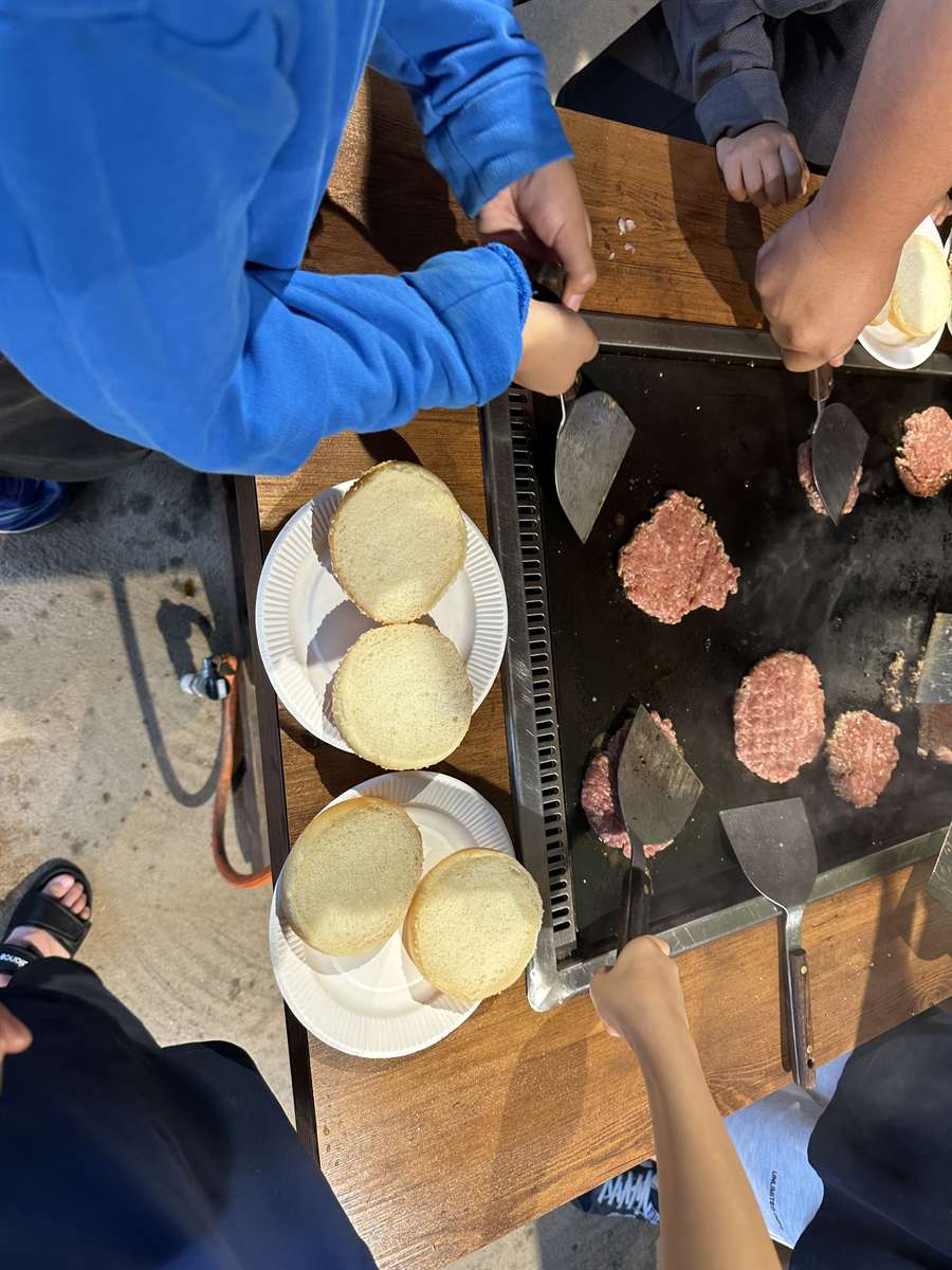屋外の鉄板でハンバーガーのパティを焼く様子。バンズが紙皿に並ぶ
