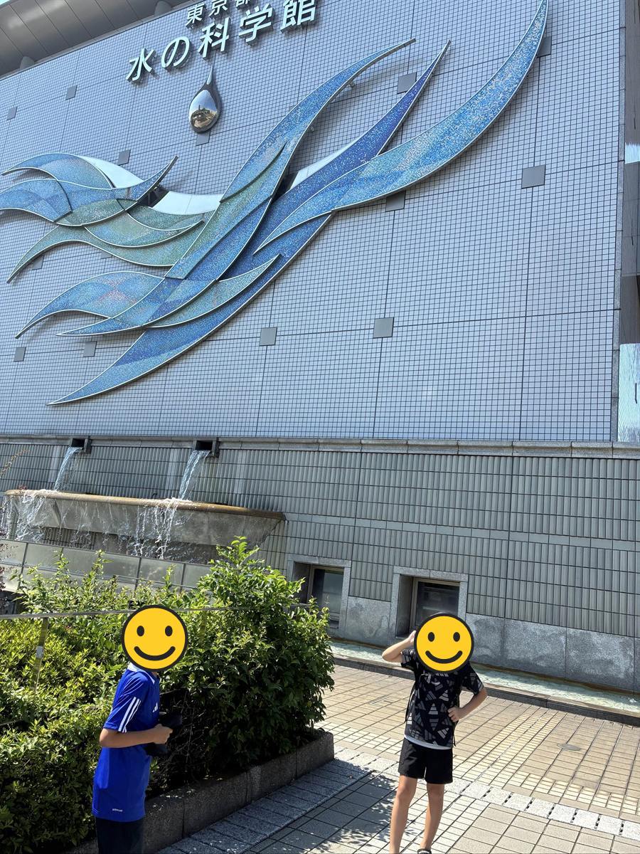 東京都水の科学館の外壁と青い波のオブジェの前に立つ子ども2人