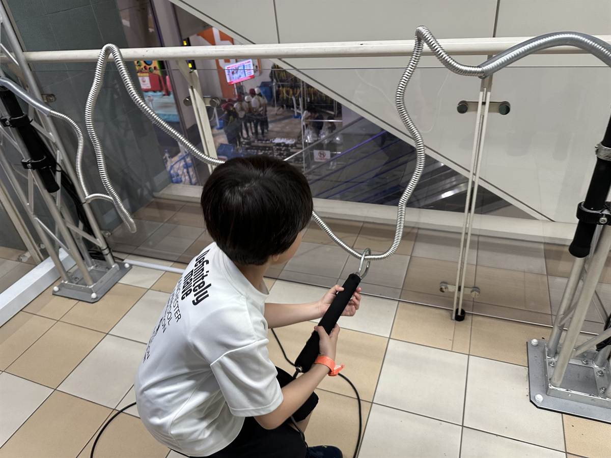曲がりくねった金属のレールに沿って輪を動かす、電流イライラ棒のような遊具に挑む子ども