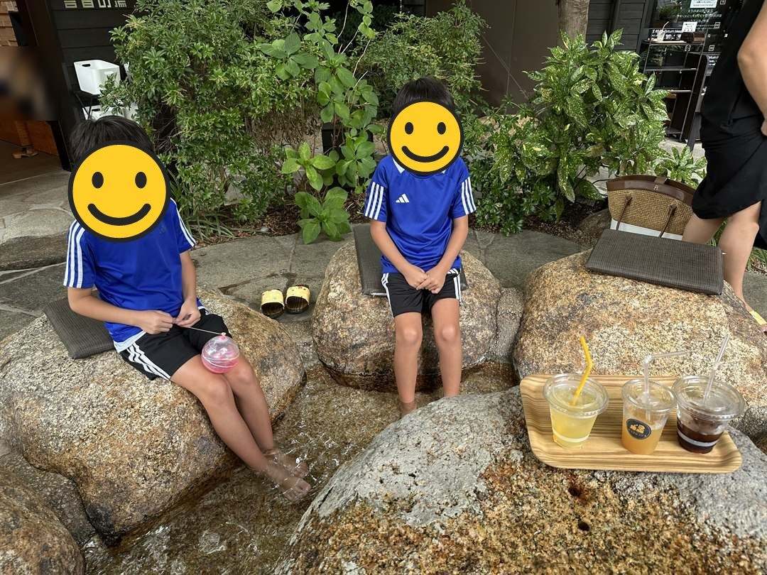 狐の足あとの屋外の足湯で岩に座って足をつける兄弟と、カフェのドリンク