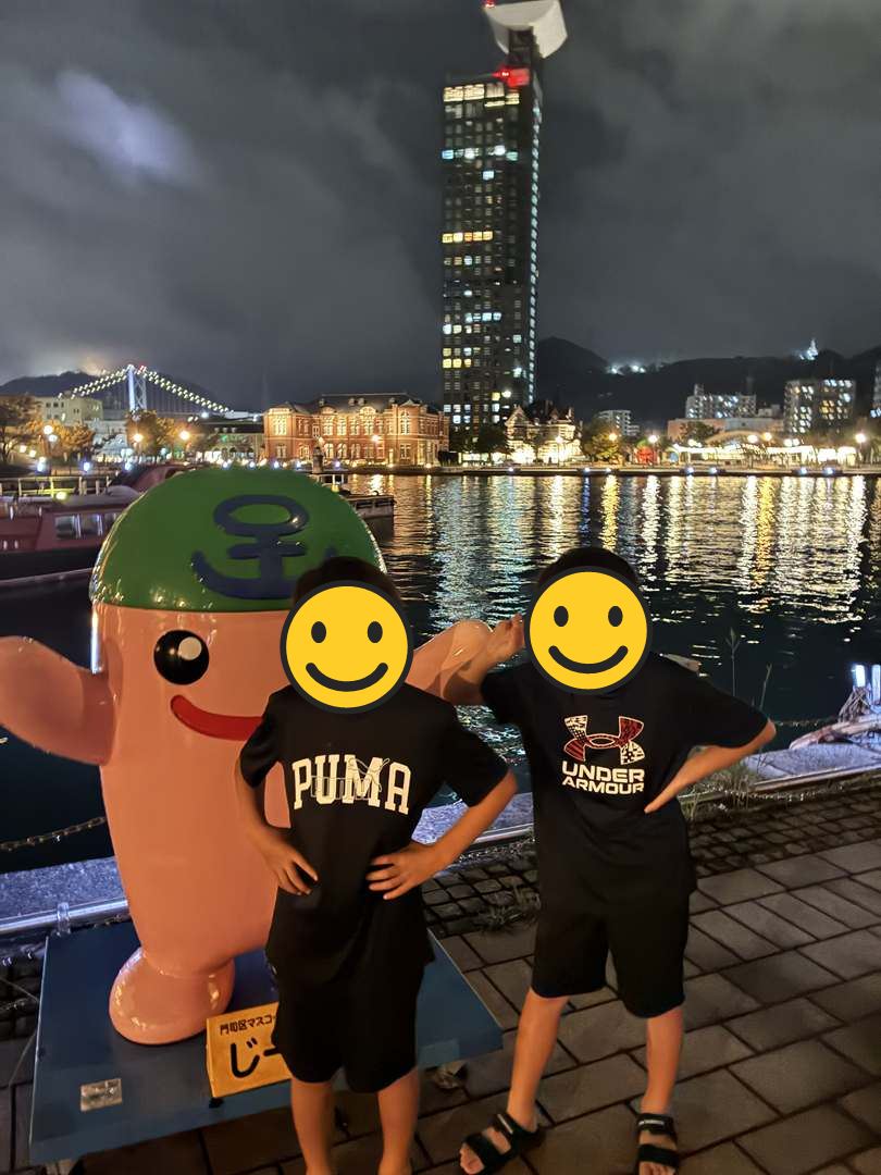 夜の門司港レトロで門司区のマスコットの像の横に立つ兄弟。背景にライトアップされた関門橋