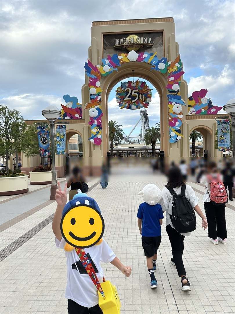 USJの25周年の飾りがついた入場ゲートの前で、かぶりものをしてピースするこども