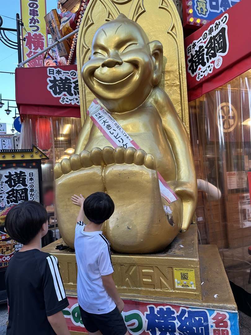 串かつ横綱の店先の大きな金色のビリケンさんの足の裏をなでる兄弟の後ろ姿